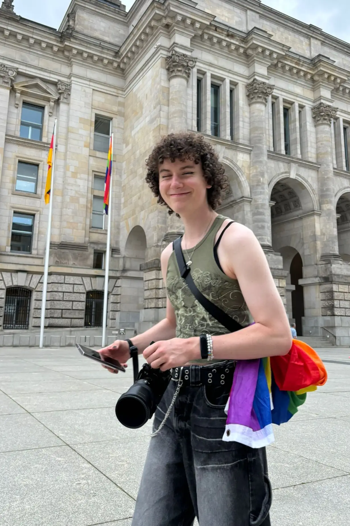 The picture shows the Owner of the Website, Melly, holding her Camera and smiling. She has a pride flag around her waist, as she is part of the Pride Parade on this picture.